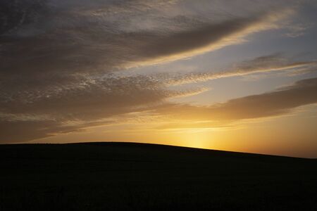 Mysterious dark landscape with the silhouette of a hill against yellow sunlight and clouds on the sky at duskの写真素材