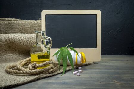 Marijuana and drug abuse or addiction still life with a fresh green leaf draped over a bottle of tablets in front of a blank slate with copy space, coiled rope and decanter of oil on a rustic tableの写真素材