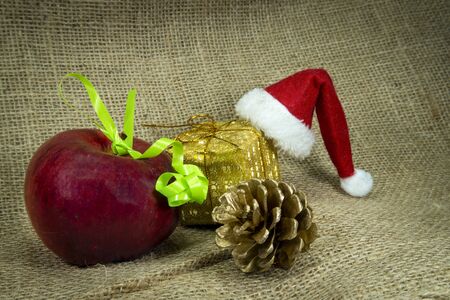 Christmas still life with colorful red Santa Hat, decorations, gold wrapped gift and apple over a hessian or burlap background with copy space for a seasonal greetingの写真素材