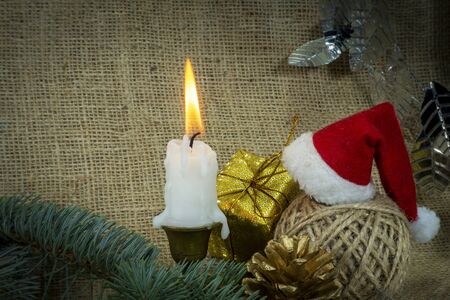 Christmas background with burning candle, decorations, gift, ball of twine and Santa hat on rustic burlap background with copy space for a holiday greetingの写真素材