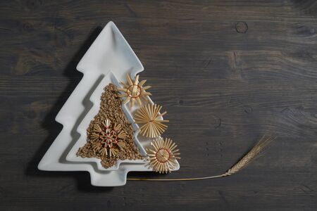 Two decorative Christmas tree shape plates lying on top of each other decorated with natural seeds, handcrafted straw decorations and ear of wheat over a gray weathered wood background with copy spaceの写真素材