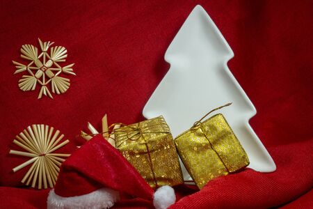 Festive red Christmas still life with gifts, handmade straw snow flakes, white Xmas tree and a Santa hat over colorful bright red fabric to celebrate the holiday season, free copy spaceの写真素材