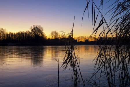 Reeds on a tranquil frozen lake at sunset with colorful reflections and a vivid orange skyの写真素材