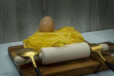 Homemade Italian ribbon pasta still life with a plate of cooked pasta, whole fresh egg, wooden rolling pin and gold colored eating utensilsの写真素材