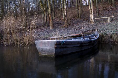 Old wooden dinghy moored to the shore of a lake in winter with woodland trees, an empty rustic bench and reflections on the waterの写真素材