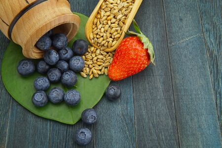 Food still life with fresh ripe blueberries spilling onto a green leaf and a strawberry flanking a wooden scoop with raw wheat seeds on a black wood background with copy spaceの写真素材