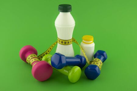 Weight loss and fitness concept with tape measure wound round a white plastic bottle with a slim line shape, pink and blue dumbbells on green chroma key backgroundの写真素材