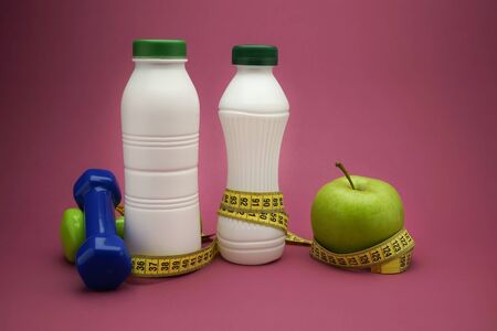 Healthy diet and weight loss concept with dumbbells, a tape measure wound round a fresh green apple and two white plastic bottles, one with a slim line shape, over a pink backgroundの写真素材