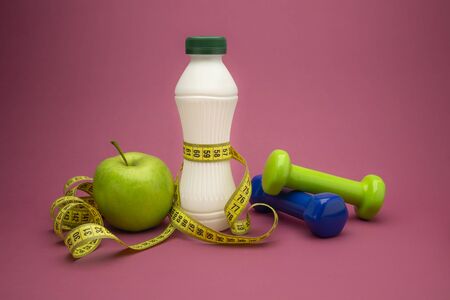 Slimming and weight loss concept with green apple, dumbbells and tape measure wound round a white plastic bottle with a slim line shape, over a pink backgroundの写真素材