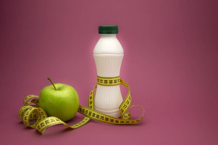 Slimming and weight loss concept with green apple, a tape measure wound round a white plastic bottle with a slim line shape, over a pink backgroundの写真素材