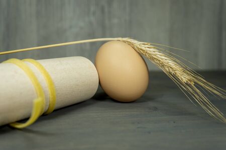 Italian ribbon pasta wound round a wooden rolling pin with whole egg and ear of golden wheat in a conceptual food still lifeの写真素材