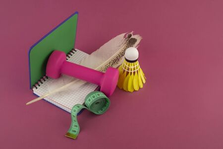 Sport weight loss and fitness concept with a shuttlecock, dumbbell weight and tape measure alongside an open notebook with quill pen on a bright pink backgroundの写真素材