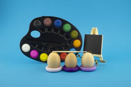 Easter egg decoration concept with paint brushes, blank blackboard and set of watercolor paints in pallet. Studio still life on blue backgroundの写真素材