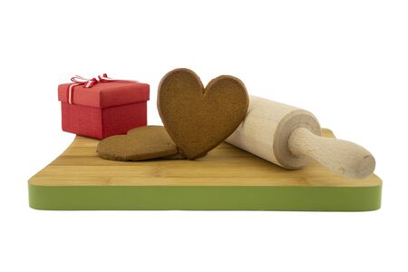 Heart-shaped cookies gift preparation concept with brown biscuits next to a gift box and rolling pin on wooden cutting board isolated on white backgroundの写真素材
