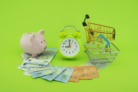 Cost of shopping or spending savings concept with banknotes scattered on a green background with a piggy bank, clock, small wire shopping basket and cartの写真素材