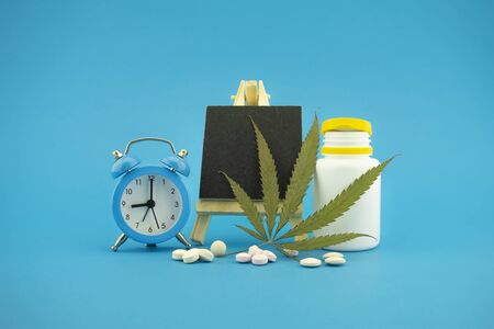Medicinal marijuana concept with fresh green leaf, pharmaceutical bottle and pills, alarm clock and small chalk board over a blue background with copy spaceの写真素材