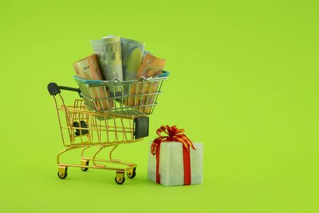 Miniature shopping cart filled with cash banknotes and a small gift box isolated on green background. Saving money for spending on gifts conceptの写真素材