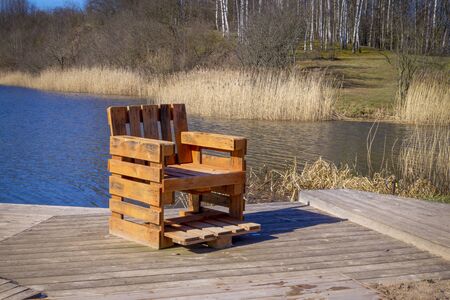 Rustic hand crafted chair made from wood pallets on wooden deck overlooking a river or lake with reeds in spring sunshineの写真素材