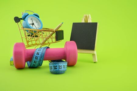 Going shopping for sports diet concept still life with dumbbells, measuring tape, blank blackboard, alarm clock and wire shopping cart on green backgroundの写真素材