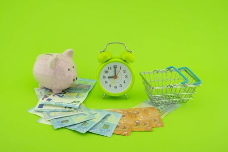 Cost of shopping or spending savings concept with banknotes scattered on a green background with a piggy bank, clock and small wire shopping basketの写真素材
