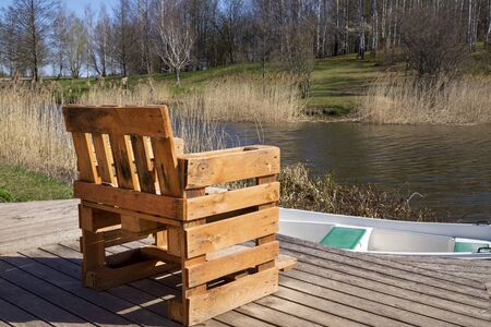 Rustic hand crafted chair made from wood pallets on wooden deck overlooking a river or lake with reeds in spring sunshineの写真素材