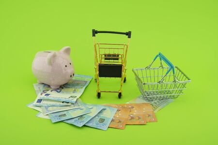 Cost of shopping or spending savings concept with banknotes scattered on a green background with a piggy bank, small wire shopping basket and cartの写真素材