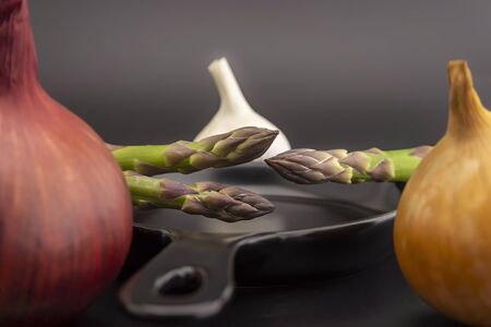 Fresh seasonal spring cooking ingredients surrounding a cast iron skillet with red and white onions, garlic bulb and green asparagus spears in a low angle viewの写真素材
