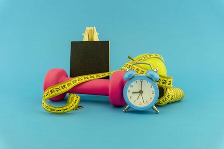 Starting healthy lifestyle concept. Still life with pink dumbbell, measuring tape, apple, chalkboard and alarm clock on blue backgroundの写真素材
