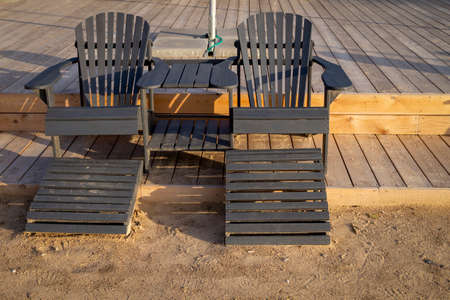 Two empty wooden beach chairs at the edge of a deck in warm evening light in a summer vacation and...