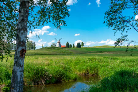 Lush green landscape with windmill and stream meandering through trees under a sunny blue sky in spring or summerの写真素材