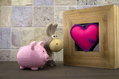 Cute little piggy bank and toy donkey with a handcrafted pink heart in a wooden frame against a tiled backgroundの写真素材