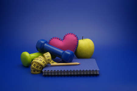Fitness and weight loss concept with dumbbells, measuring tape, heart, green apple and new notebook on blue backgroundの写真素材