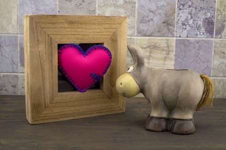 Cute little toy donkey look at handcrafted pink heart in a wooden frame against a tiled backgroundの写真素材