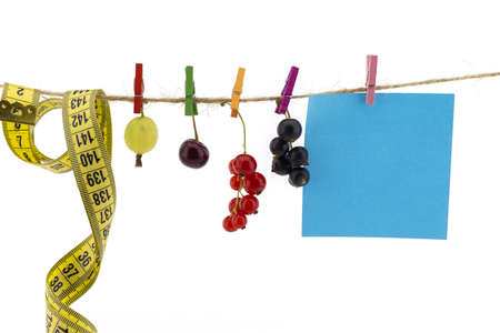 Fresh fruit, tape measure and blank sticky note hanging from clothes pegs on a rope with a gooseberry, red currants, cherry and black currants in a healthy diet conceptの写真素材