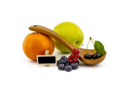 Fresh seasonal fruit still life with small signboard, assorted berries including blueberries, cherries and red currants on wooden spoons with apple and orange on a white backgroundの写真素材