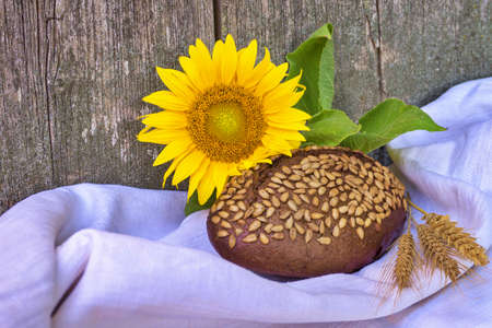 Healthy loaf of brown wholewheat bread coated in sunflower seeds with colorful fresh yellow sunflower and golden ears of wheat in a food still lifeの写真素材