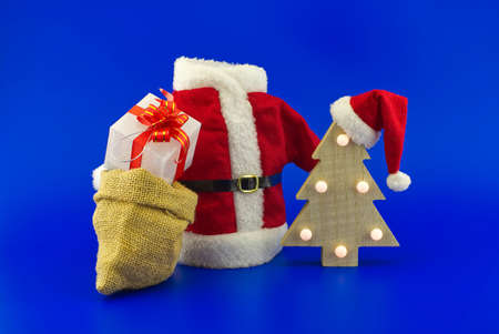 Red Santa Claus suit near stylized wooden Christmas tree with Santa hat and jute sack with gift boxes on a festive blue background. New Year and Christmas gift season conceptの写真素材