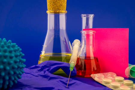 Concept of research for a virus vaccine with a conical lab flasks with yellow and red solutions alongside a hypodermic syringe and blue virus molecules with blank square of paper note for textの写真素材