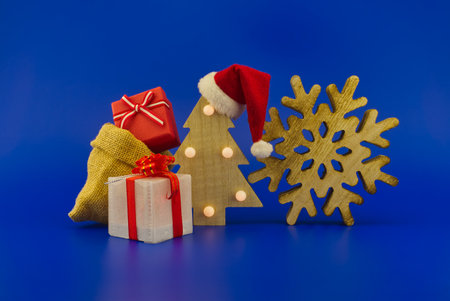 Stylized wooden Christmas tree with red Santa hat and jute sack with gift boxes on a festive blue background. New Year and Christmas gift season conceptの写真素材