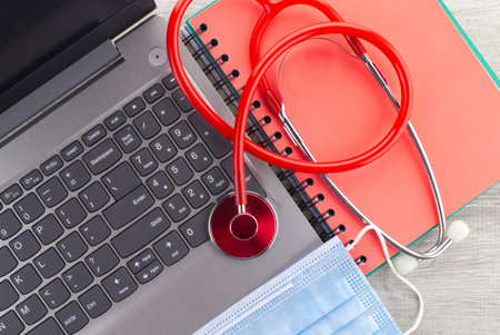 Coronavirus or Covid-19 medical concept with a colorful red stethoscope coiled over a surgical face mask alongside a notebook and pen on a wooden desk with computer keyboard viewed top downの写真素材