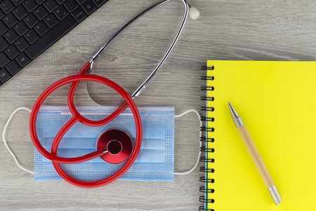 Coronavirus or Covid-19 medical concept with a colorful red stethoscope coiled over a surgical face mask alongside a notebook and pen on a wooden desk with computer keyboard viewed top downの写真素材
