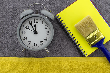 Color of the year and color trends themed still life with alarm clock placed to the side of yellow wire bound notepad and paintbrush on a textured grey background viewed as a flat lay from overheadの写真素材
