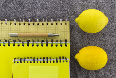 Colorful yellow themed still life with fresh healthy lemons placed to the side of three wire bound notepads and pen on a textured grey background viewed as a flat lay from overheadの写真素材