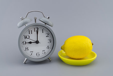 Gray alarm clock and lemon on a yellow plate on grey with copy space. Color of the year and color trends conceptの写真素材