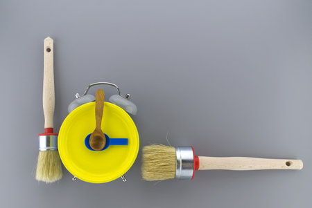 Creative DIY, decorating or renovations background with two clean bristle brushes and a colorful yellow alarm clock with tools for hands forming a border on grey with copy spaceの写真素材