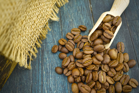 Roasted coffee beans spilling from a wooden scoop on wooden and hessian or burlap fabric backgroundの写真素材