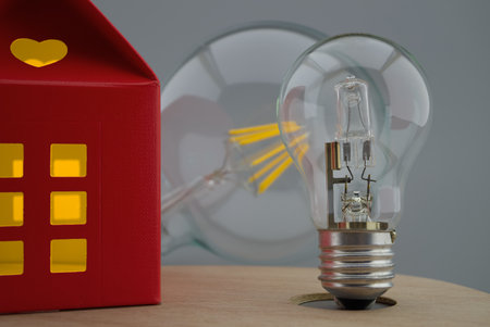 Incandescent lamp light bulbs next to a small red house of paper with a cut out heart and warm light in windows. Close-up concept of home lightingの写真素材