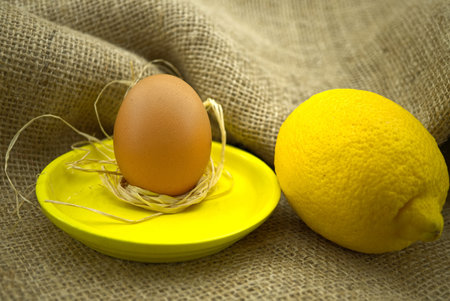 Brown egg on bright yellow plate near yellow lemon over rustic hessian sack background. Organic farming and color trends concept with copy spaceの写真素材