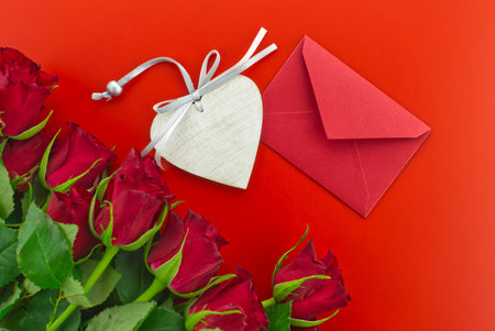Red roses, envelope and wooden heart medallion on red background with free copy space. Wishes, greetings and love messageの写真素材