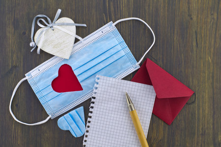 Face mask with cut out heart shape alongside a wire bound notepad and pen and red envelope on wooden background with free copy space. Social distancing and contact-less greetingsの写真素材
